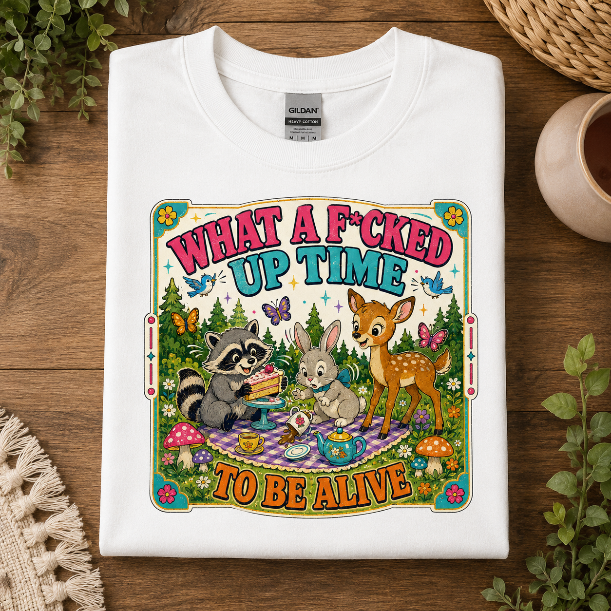 A folded white Gildan t-shirt with a colorful graphic print, displayed on a wooden surface with decorative plants and a woven cup.
