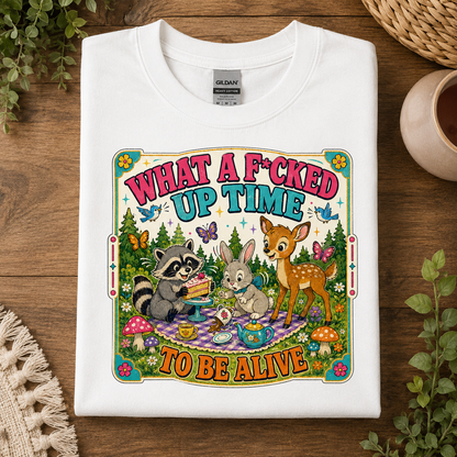 A folded white Gildan t-shirt with a colorful graphic print, displayed on a wooden surface with decorative plants and a woven cup.
