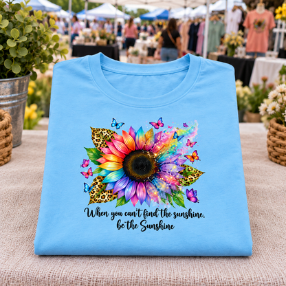 A sky blue folded T-shirt displayed on a table at an outdoor market. The shirt features a large, vibrant rainbow sunflower graphic with colorful butterflies and leopard-print leaves, plus the text 'When you can't find the sunshine, be the Sunshine' in black script.