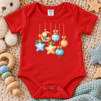A red short-sleeve baby bodysuit laid flat on a beige textured blanket with soft toys around it. The bodysuit has a colorful Christmas ornament graphic on the chest, including a crescent moon, stars, and hanging ornaments.