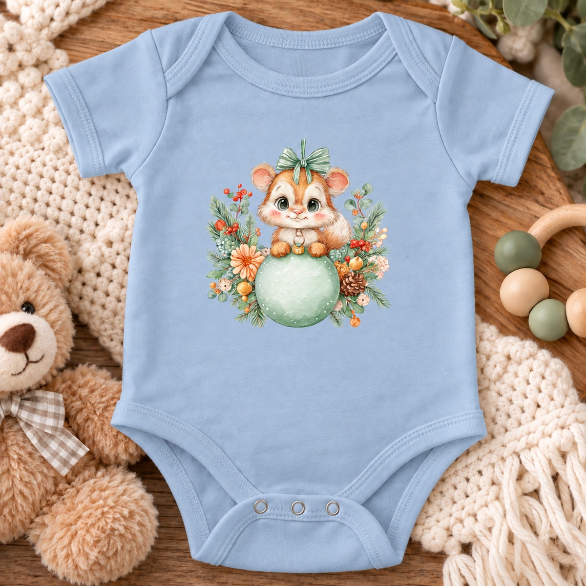 A light blue baby bodysuit laid flat on a wooden surface, featuring a cute cartoon chipmunk sitting on a green ball surrounded by autumn leaves and flowers. The bodysuit is styled with a plush teddy bear, a cream knit blanket, and wooden baby toys.