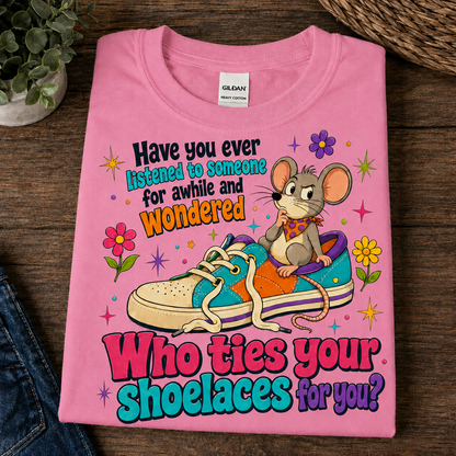 A folded azalea t-shirt with a humorous graphic print featuring a cartoon mouse sitting on a sneaker and the text 'Have you ever listened to someone for awhile and wondered Who ties your shoelaces for you?', displayed on a dark wood background.