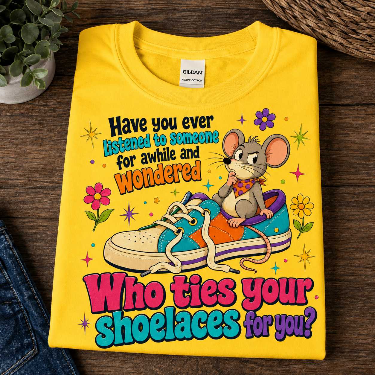 A folded daisy colored Gildan t-shirt with a colorful graphic of a mouse sitting on a sneaker and the text 'Have you ever listened to someone for awhile and wondered Who ties your shoelaces for you?', displayed on a dark wood background.