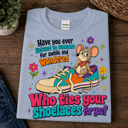 A folded light blue Gildan t-shirt with a colorful graphic print featuring a cartoon mouse sitting on a sneaker and the text 'Have you ever listened to someone for awhile and wondered Who ties your shoelaces for you?', displayed on a dark wood background.