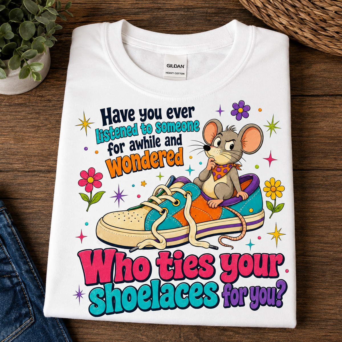 A folded white Gildan t-shirt with a colorful graphic print featuring a cartoon mouse sitting in a sneaker and the text 'Have you ever listened to someone for awhile and wondered Who ties your shoelaces for you?', displayed on a dark wood background.