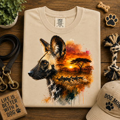 A folded sand stone colored T-shirt displayed on a wooden surface with dog-themed props. The shirt features a large watercolor-style graphic of a wild dog in profile, with an African savanna sunset scene inside the head shape.
