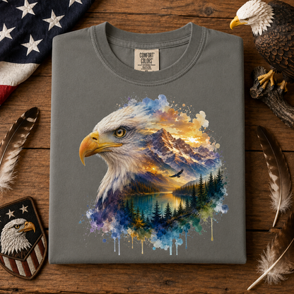 A folded gray t-shirt with a detailed graphic of an eagle head merging into a watercolor landscape of mountains, forest, and lake. The shirt is displayed on a wooden surface surrounded by feathers, a US flag, a patch, and a real eagle figurine.