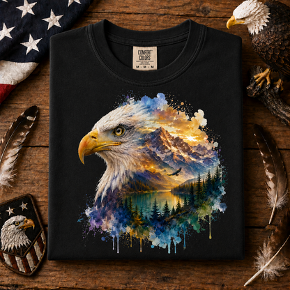 A black Comfort Colors t-shirt laid flat on a rustic wooden surface, featuring a large graphic of a bald eagle head with a colorful landscape of mountains, lake, and trees inside the feathers. The shirt is styled with patriotic props including a US flag, feathers, a patch, and a bald eagle figurine.