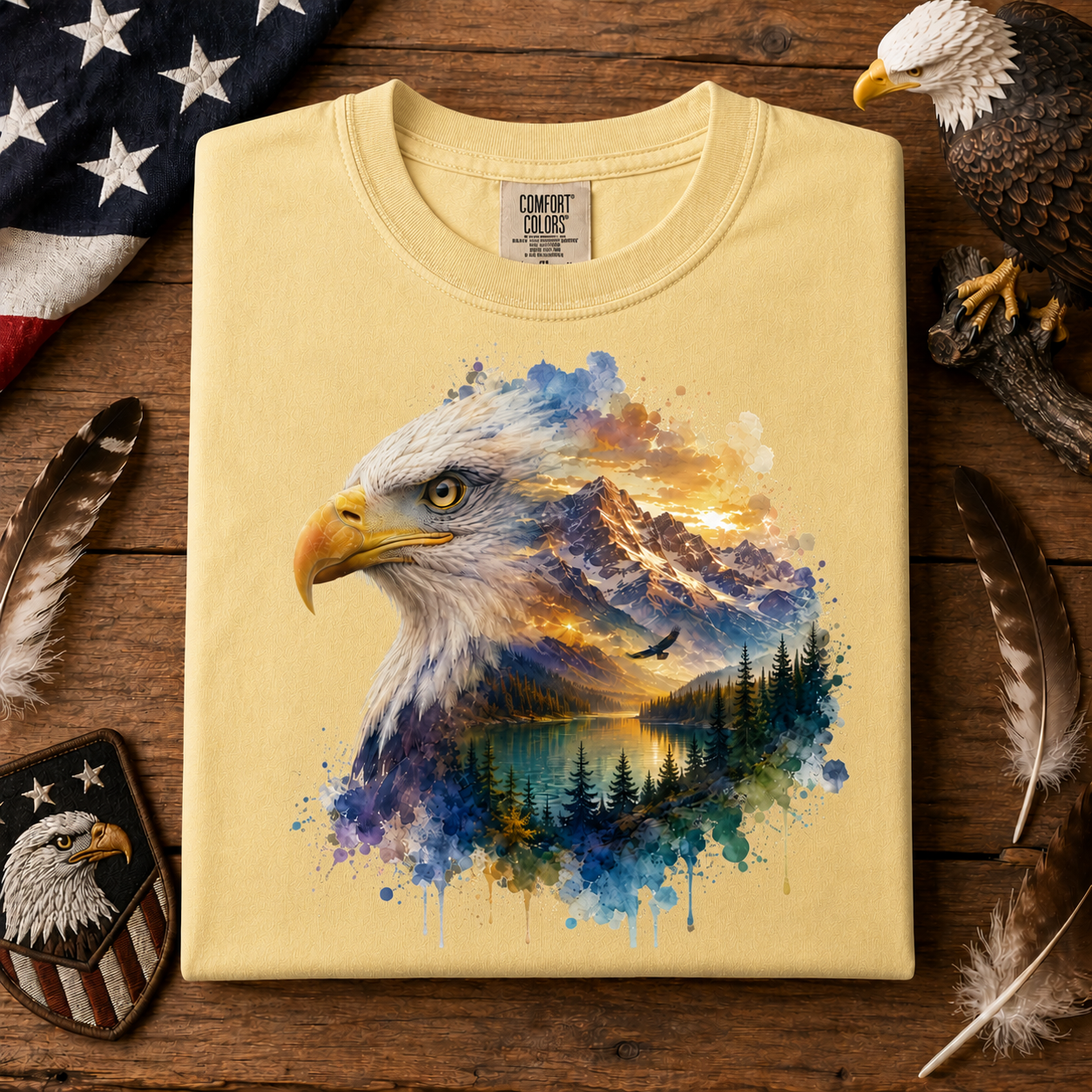 A folded butter colored T-shirt with a large colorful bald eagle and mountain landscape graphic on the front, displayed on a rustic wooden surface with feathers, a patch, and part of a flag around it.
