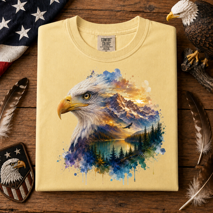 A folded butter colored T-shirt with a large colorful bald eagle and mountain landscape graphic on the front, displayed on a rustic wooden surface with feathers, a patch, and part of a flag around it.