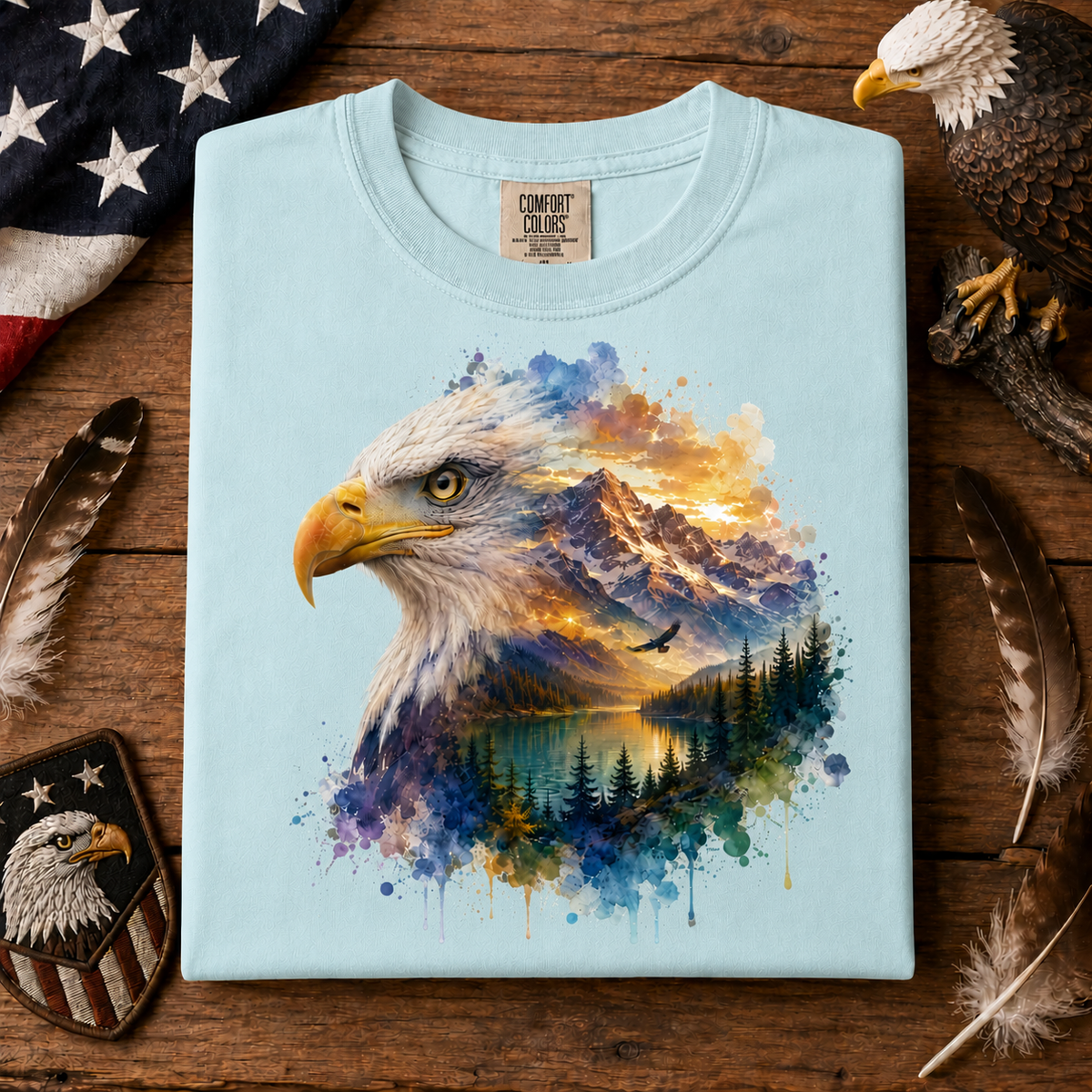 A chambray colored t-shirt with a large graphic of a bald eagle head merging into a colorful landscape of mountains, a lake, and a forest. The shirt is laid flat on a rustic wooden surface surrounded by feathers, a US flag, and a patch.