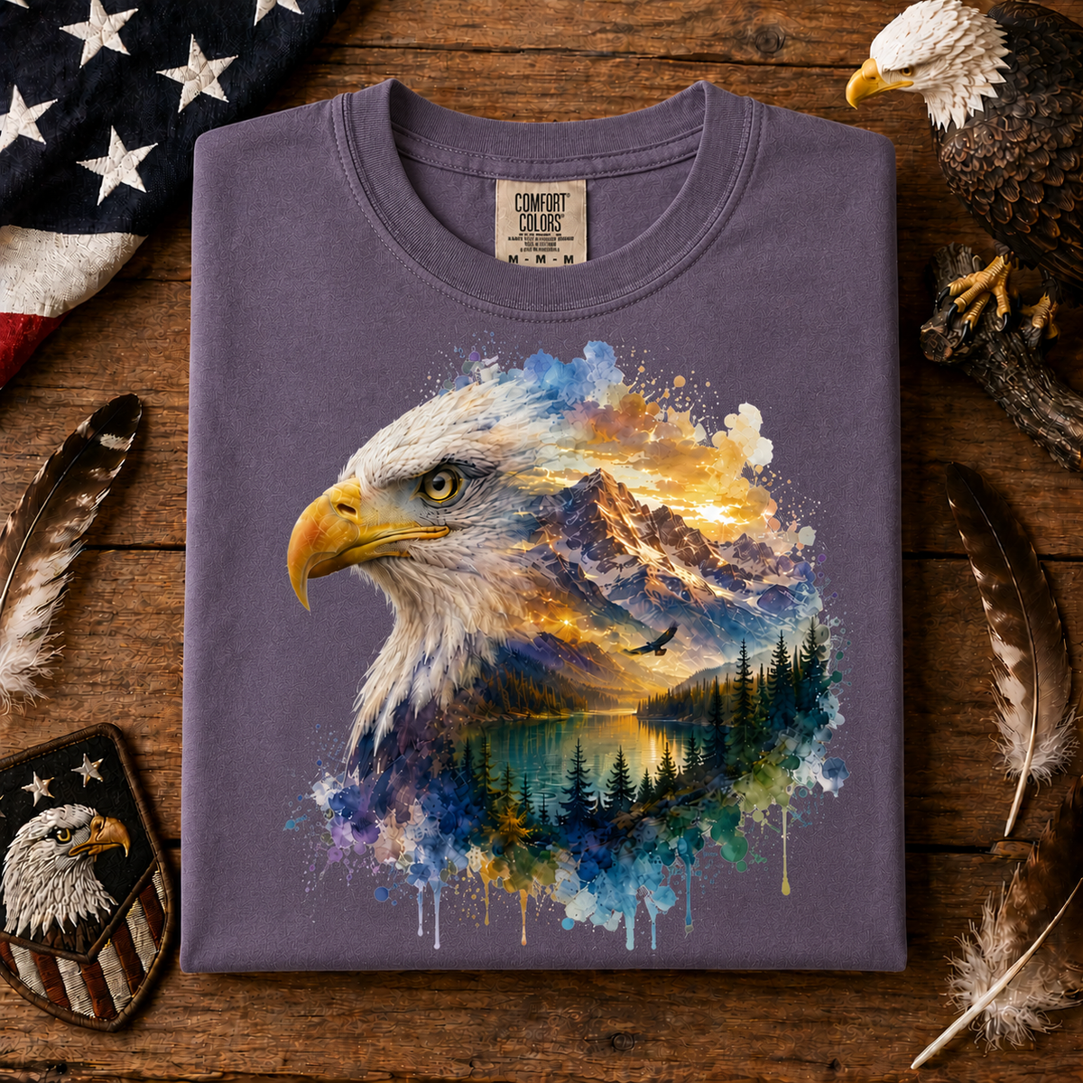 A folded wine colored t-shirt with a detailed graphic of an eagle head and a mountain lake landscape in watercolor splashes, displayed on a rustic wooden surface with patriotic props.