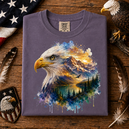 A folded wine colored t-shirt with a detailed graphic of an eagle head and a mountain lake landscape in watercolor splashes, displayed on a rustic wooden surface with patriotic props.