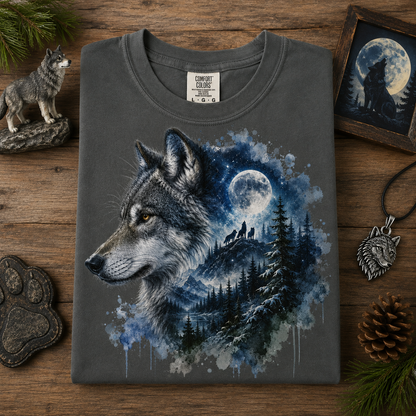 A granite gray t-shirt with a detailed graphic of a wolf in profile, set against a moonlit forest and mountain scene. The shirt is displayed on a rustic wooden surface surrounded by wolf-themed decorative props, including a small wolf figurine, a pinecone, a paw print, and a wolf pendant.