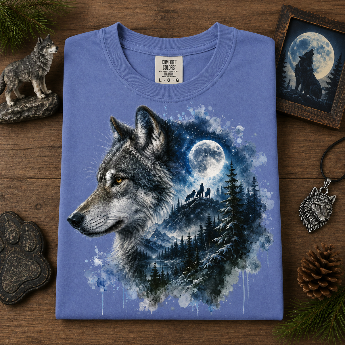 A folded periwinkle blue crewneck t-shirt with a detailed graphic of a wolf's head and a forest silhouette under a full moon, styled on a wooden surface with wolf-themed props.