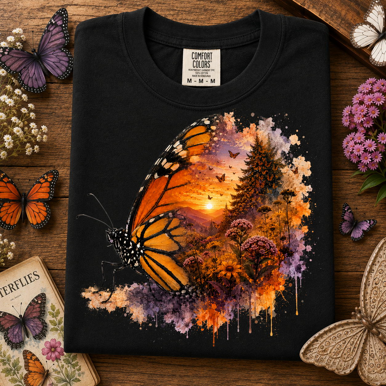 A black Comfort Colors t-shirt laid flat on a rustic wooden surface, decorated with butterflies, flowers, and a book. The shirt features a large graphic print of a monarch butterfly over a vibrant sunset landscape with trees and purple floral accents.