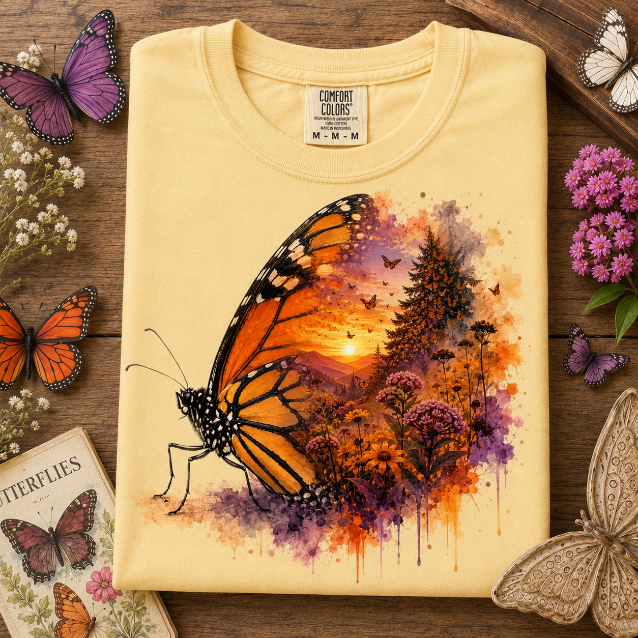 A folded butter colored t-shirt with a vibrant butterfly and landscape watercolor print, styled on a rustic wooden background with decorative butterflies and flowers.