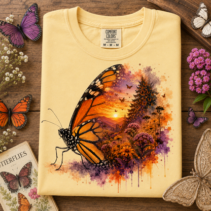 A folded butter colored t-shirt with a vibrant butterfly and landscape watercolor print, styled on a rustic wooden background with decorative butterflies and flowers.