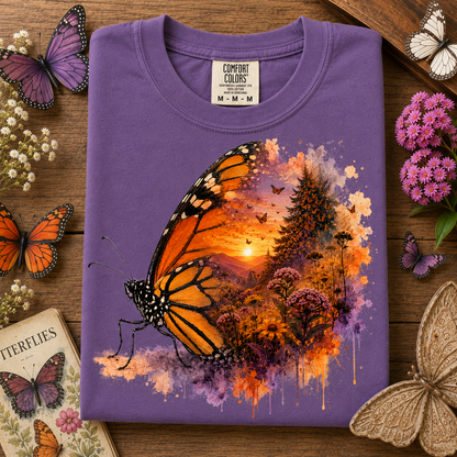 A folded grape colored t-shirt with a vibrant graphic of a monarch butterfly over a sunset forest scene, displayed on a wooden surface surrounded by decorative butterflies and flowers. The neck label reads 'Comfort Colors'.