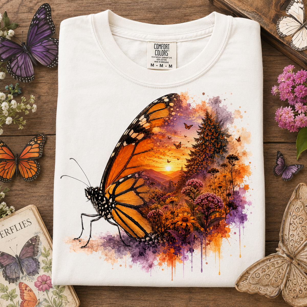 A folded white t-shirt with a vibrant butterfly and landscape graphic, displayed on a rustic wooden background surrounded by decorative butterflies and flowers. The graphic features a large orange and black monarch butterfly with a colorful watercolor sunset forest scene inside its wing.