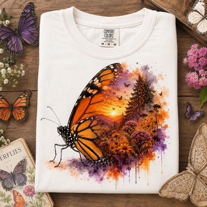 A folded white t-shirt with a vibrant butterfly and landscape graphic, displayed on a rustic wooden background surrounded by decorative butterflies and flowers. The graphic features a large orange and black monarch butterfly with a colorful watercolor sunset forest scene inside its wing.