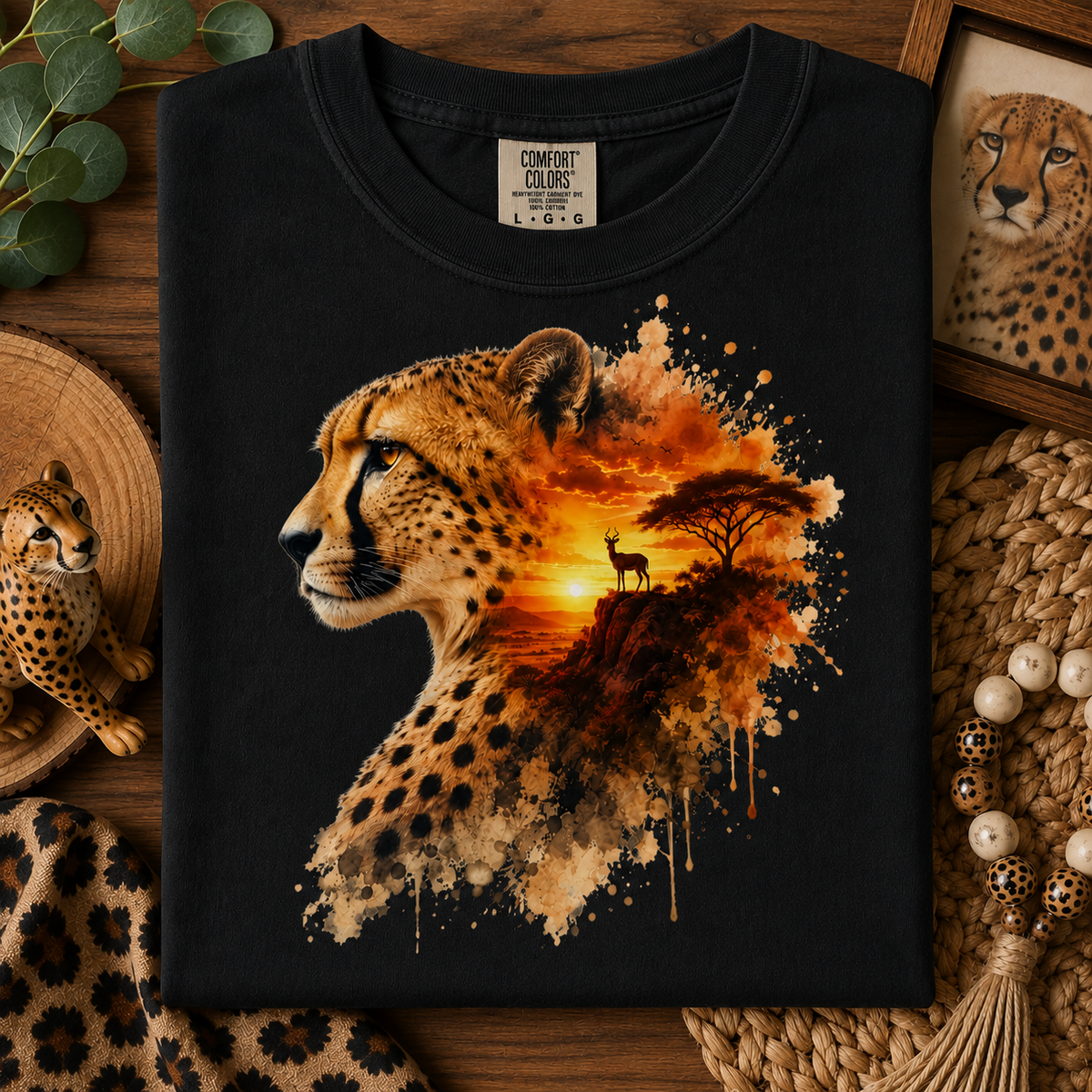 A black short-sleeve T-shirt laid flat on a rustic wooden surface, featuring a large graphic print of a cheetah profile blending into an African savanna sunset scene with a tree and bird. 