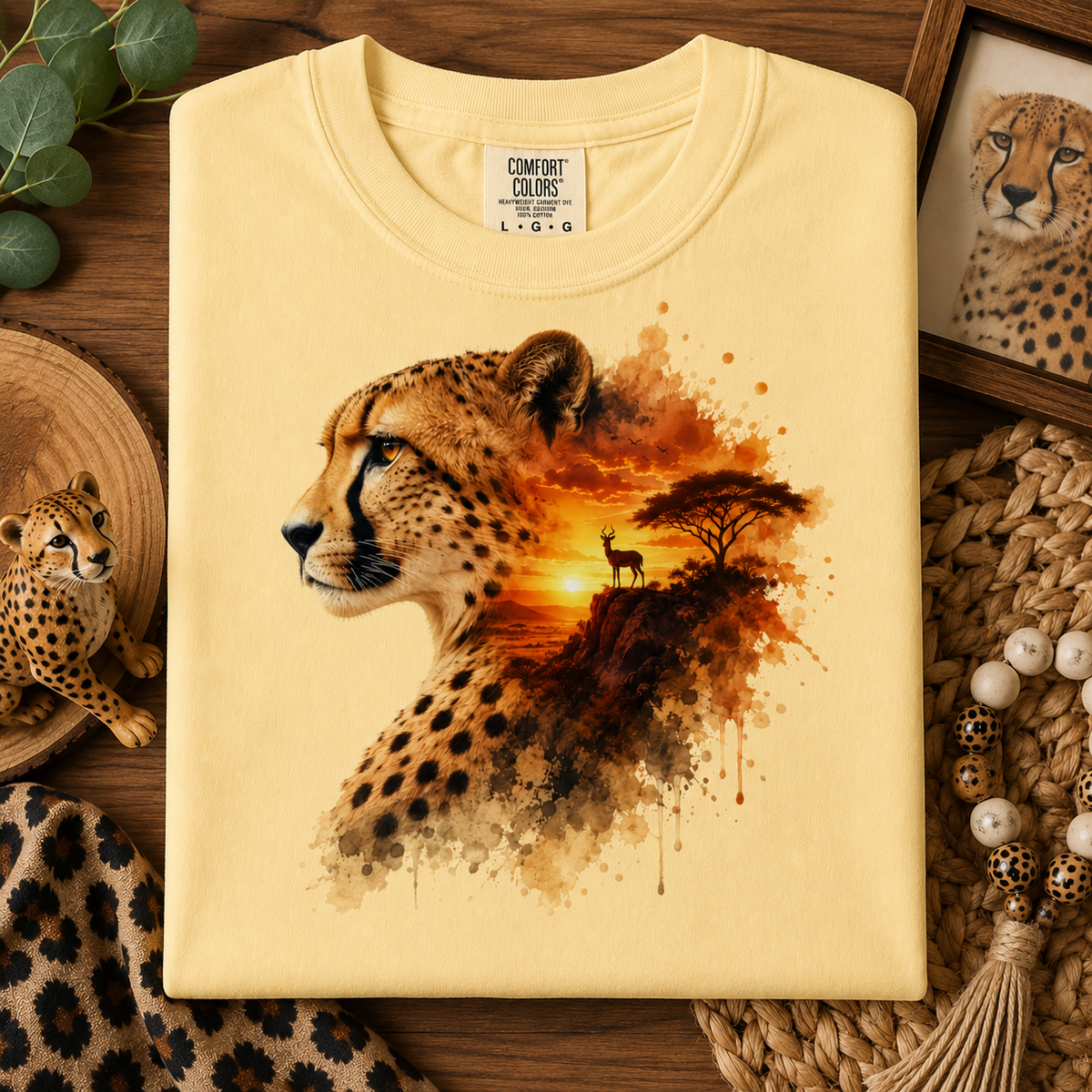 A folded butter colored t-shirt with a detailed cheetah graphic print, styled on a rustic wooden background with safari-themed props.