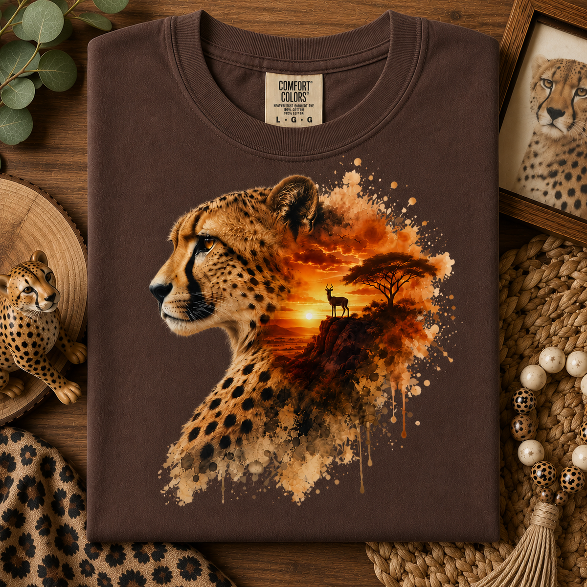 A folded expresso colored t-shirt with a detailed cheetah and savanna sunset graphic, styled in a flat-lay on a wooden surface with leopard-themed props.