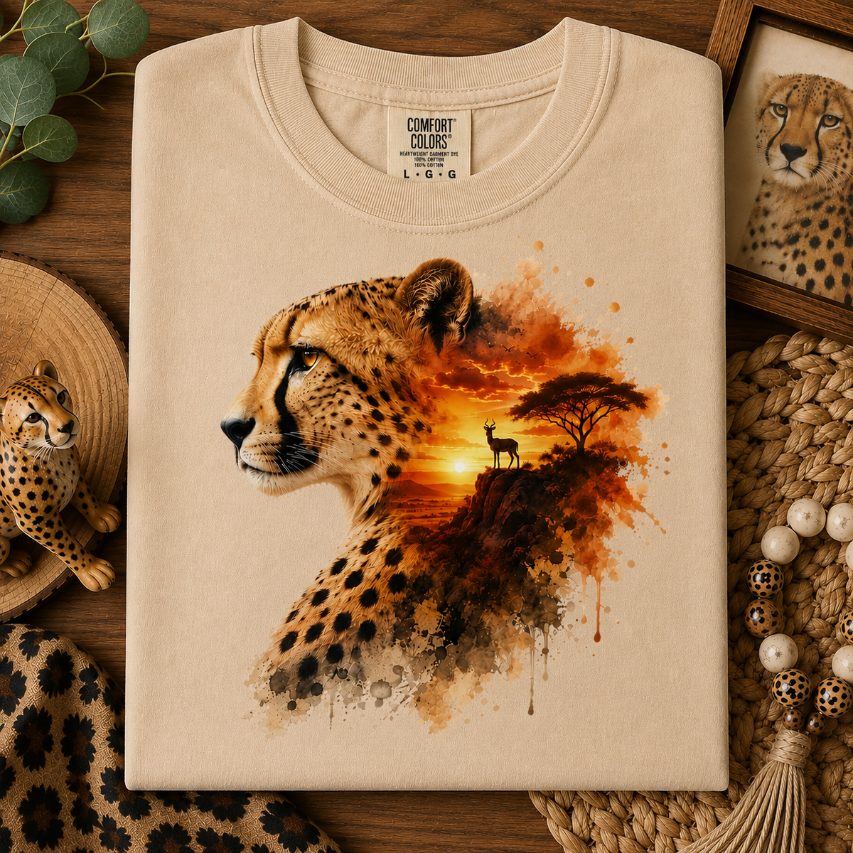 A folded sandstone colored Comfort Colors T-shirt with a detailed cheetah graphic print, styled in a flat-lay on a wooden surface with safari-themed props.