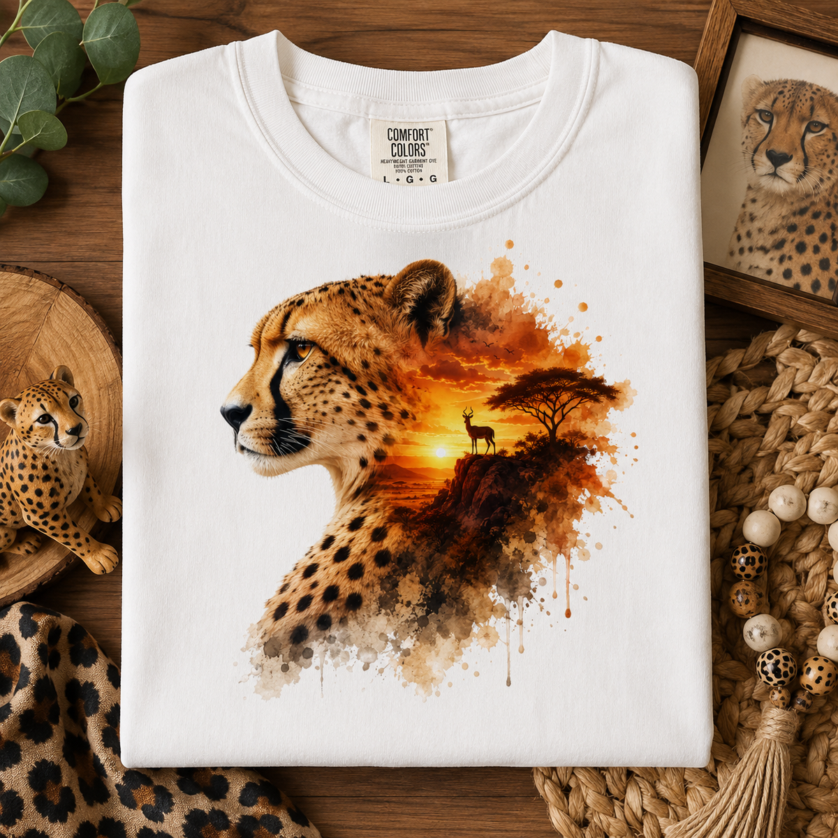 A folded white Comfort Colors T-shirt with a large cheetah graphic print, styled in a rustic flat-lay arrangement with wood, beads, and a small cheetah figurine.