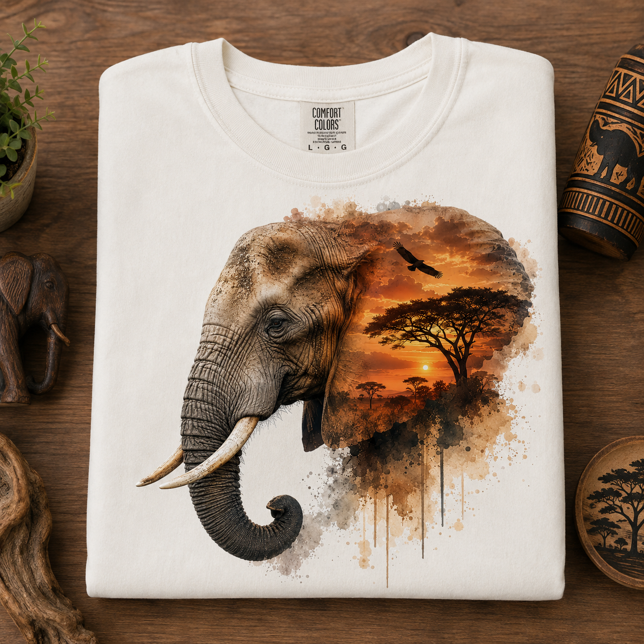 A folded white t-shirt with a graphic print of an African elephant merging into a savanna sunset scene, displayed on a rustic wooden background with decorative props.
