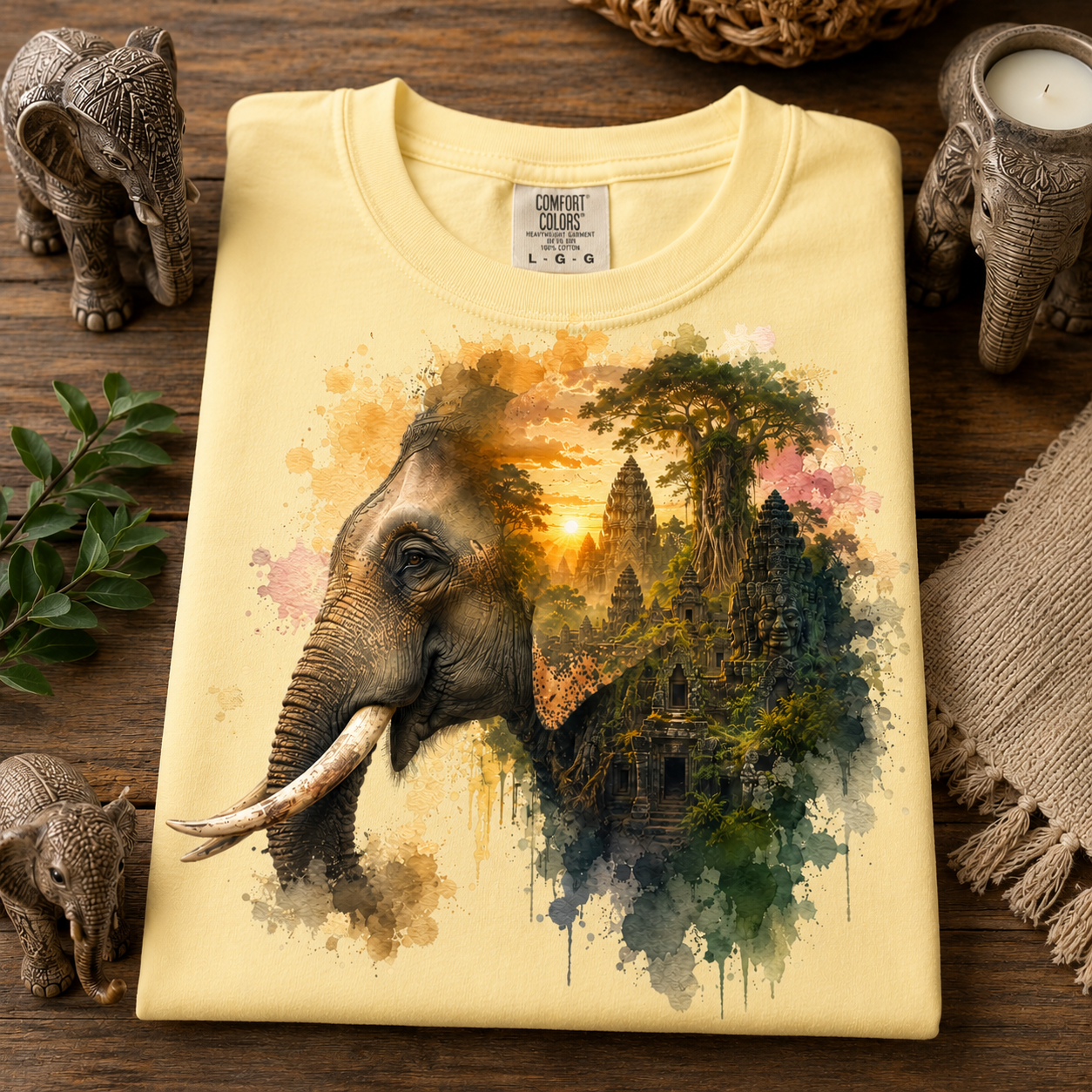A folded butter colored T-shirt is displayed on a wooden surface with elephant figurines, greenery, and woven decor around it. The shirt features a large watercolor-style graphic of an elephant merging into a jungle temple scene with a sunset.
