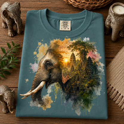 A folded emerald t-shirt with a graphic print of an elephant blending into a Cambodian temple landscape at sunset, styled on a wooden surface with elephant figurines and a woven textile.
