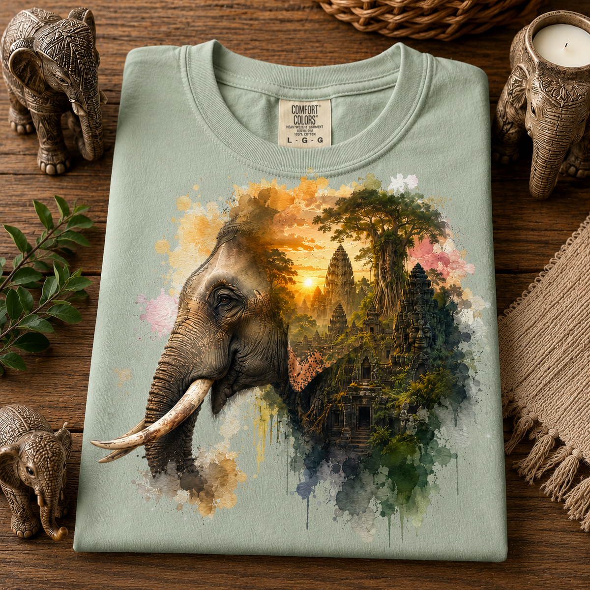 A folded hydrangea colored t-shirt with a detailed graphic print of an elephant merging into a Cambodian temple landscape at sunset, displayed on a wooden surface with decorative elephant figurines and a candle.