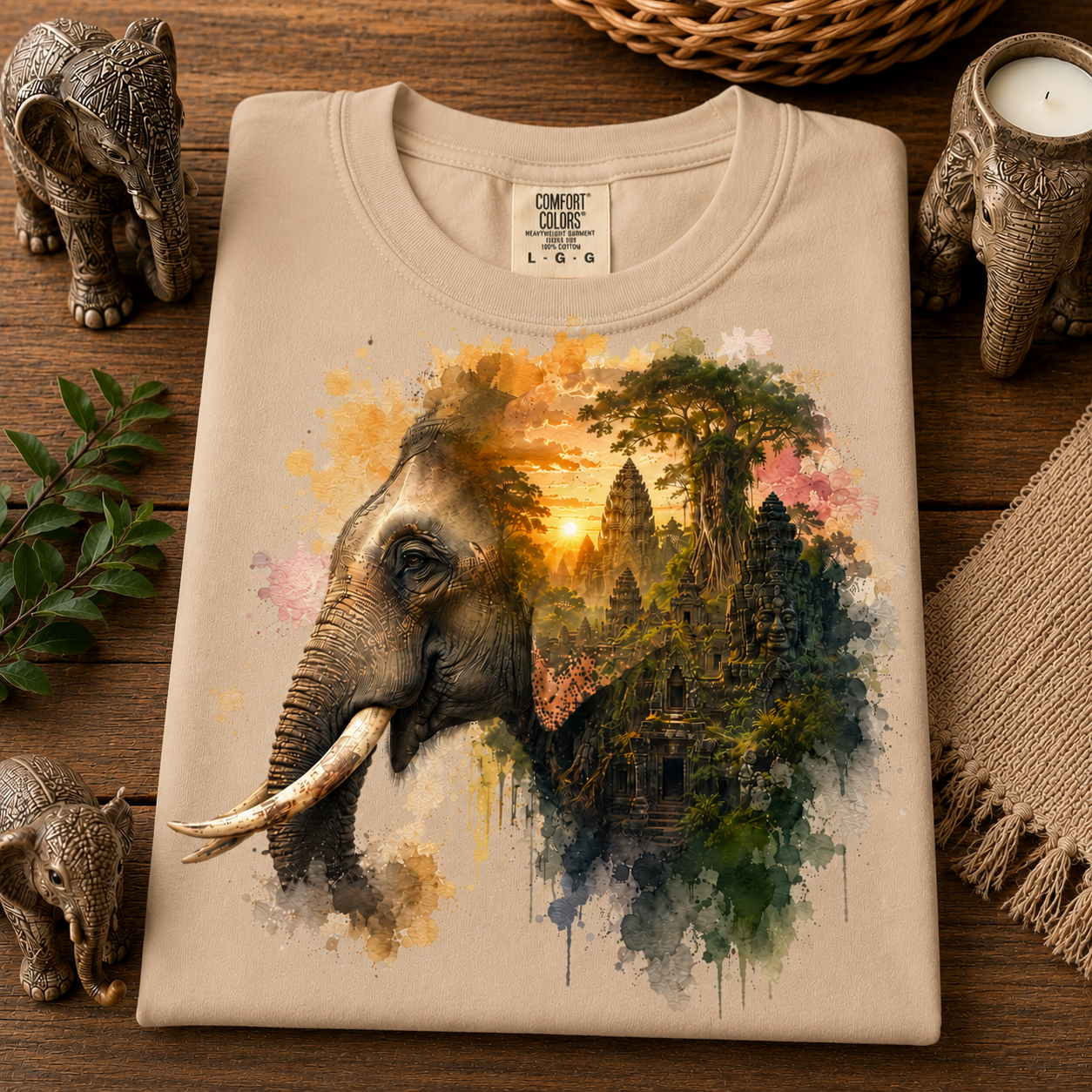 A sandstone colored Comfort Colors t-shirt is neatly folded and displayed on a rustic wooden surface. The shirt features a detailed graphic print of an elephant's head merging into a watercolor landscape of ancient temple ruins under a golden sunset.