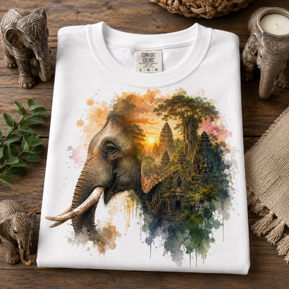 A white short-sleeve t-shirt laid flat on a rustic wooden surface, featuring a large watercolor-style graphic of an elephant merging into ancient temple ruins with a sunset sky. Decorative elephant figurines, greenery, and woven textiles surround the shirt.