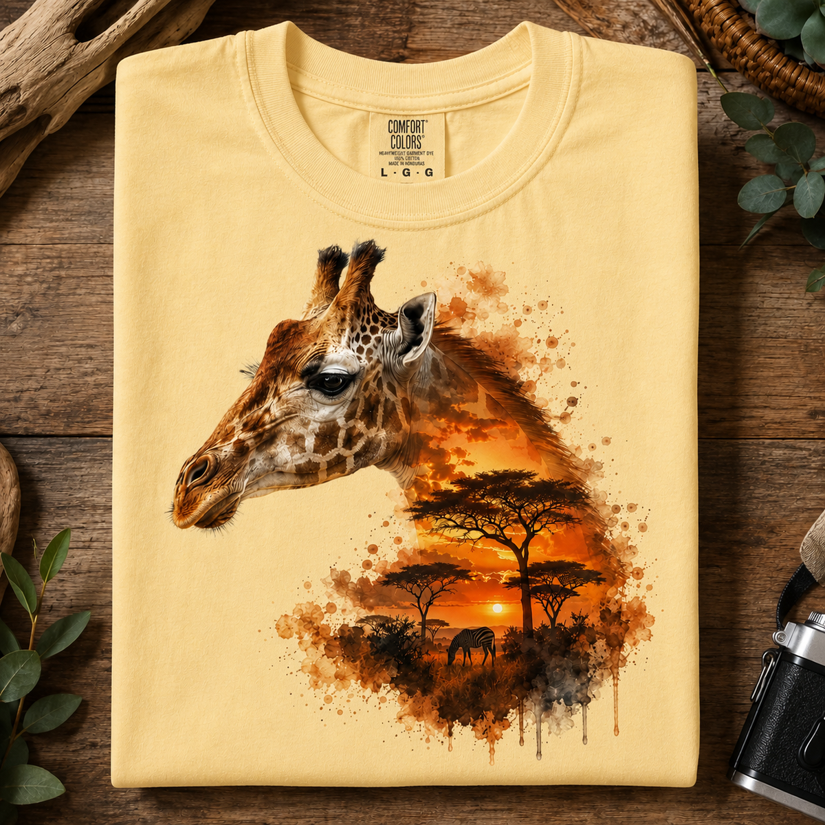 A folded butter colored t-shirt with a detailed graphic print of a giraffe head merging into an African savanna sunset scene, displayed on a rustic wooden background.