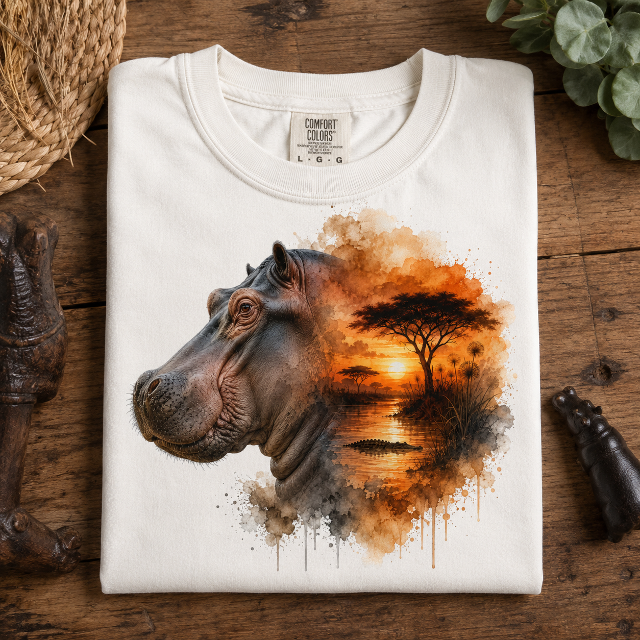 A folded white crewneck t-shirt with a graphic print of a hippopotamus head merging into an African savanna landscape, displayed on a rustic wooden surface with decorative props.