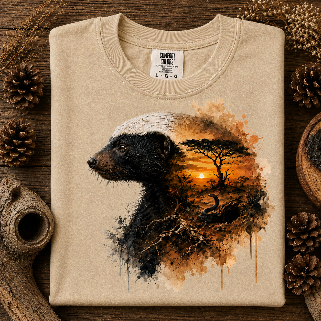 A sandstone colored crewneck sweatshirt laid flat on a rustic wooden surface, decorated with pinecones and dried grass. The front features a graphic print of a badger's head merging into an African savanna landscape with an acacia tree and sunset.