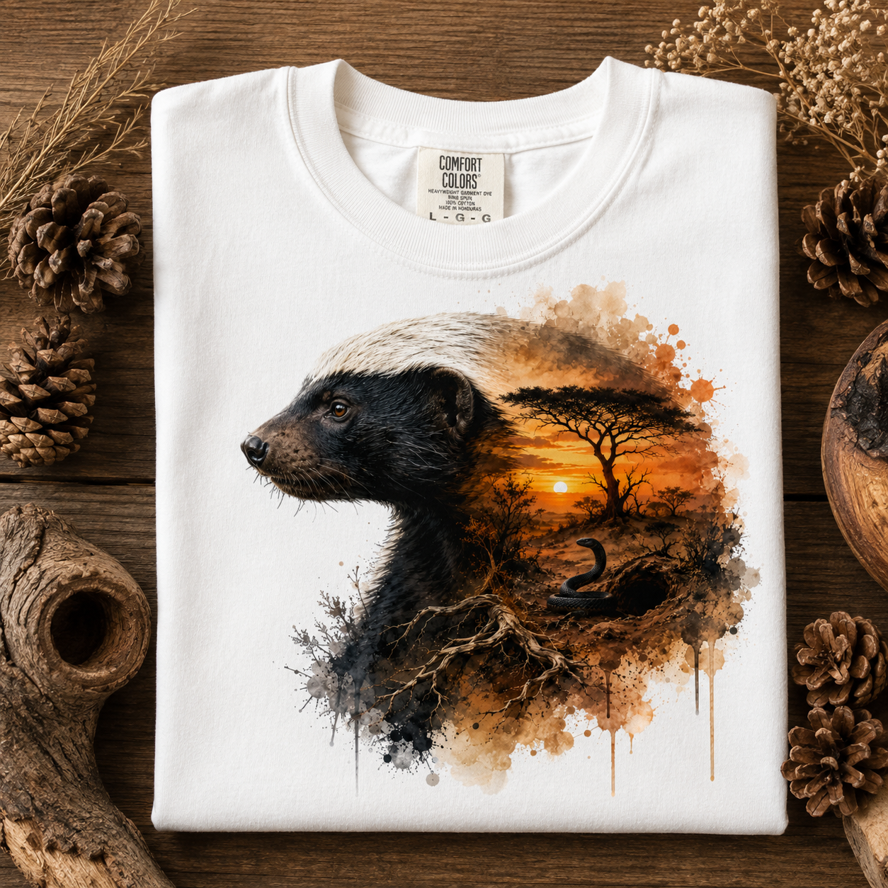 A white Comfort Colors t-shirt with a detailed graphic print of a badger and an African savanna landscape, laid flat on a rustic wooden background with pinecones and dried grass.