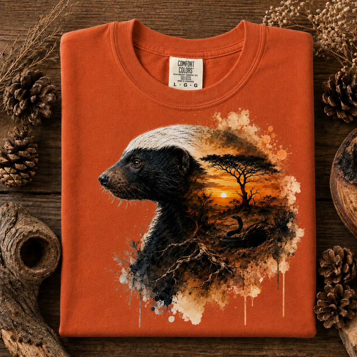 An yam colored t-shirt with a graphic print of a badger and a sunset landscape, styled on a rustic wooden background with pinecones and dried grass.