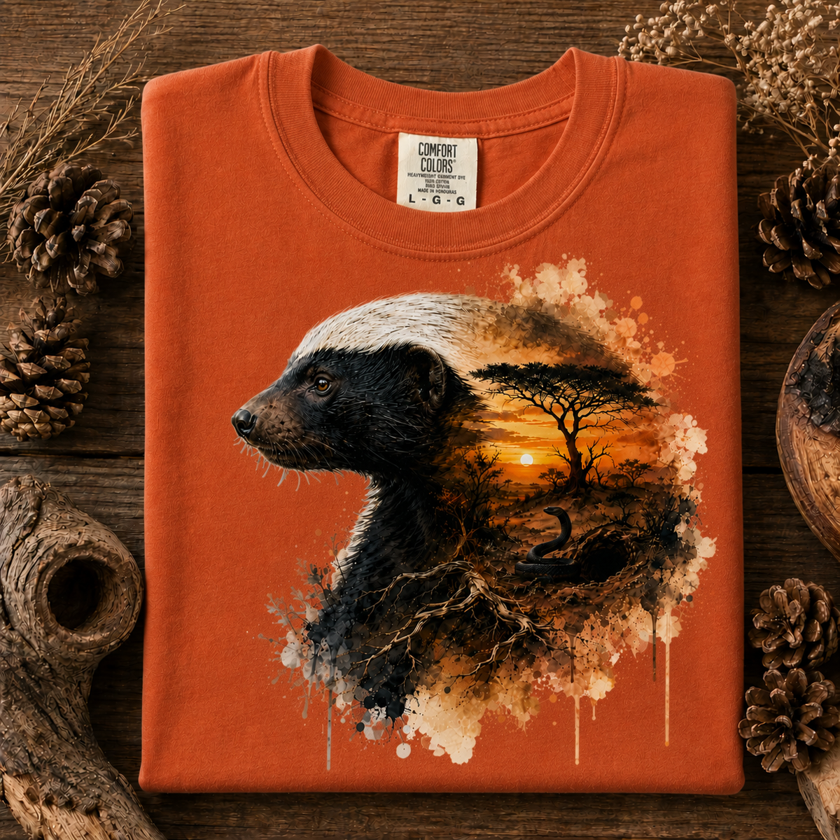 An yam colored t-shirt with a graphic print of a badger and a sunset landscape, styled on a rustic wooden background with pinecones and dried grass.