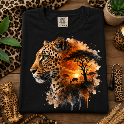 A black Comfort Colors t-shirt laid flat on a wooden surface, featuring a large graphic print of a leopard's head merging into an African savanna sunset scene. The shirt is styled with leopard-print accessories, including a small leopard figurine, a keychain, and a leopard-print fabric runner.