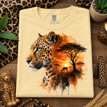 A folded butter colored t-shirt with a graphic print of a leopard's head merging into an African savanna sunset scene, styled in a flat-lay on a wooden surface with leopard-print accessories.