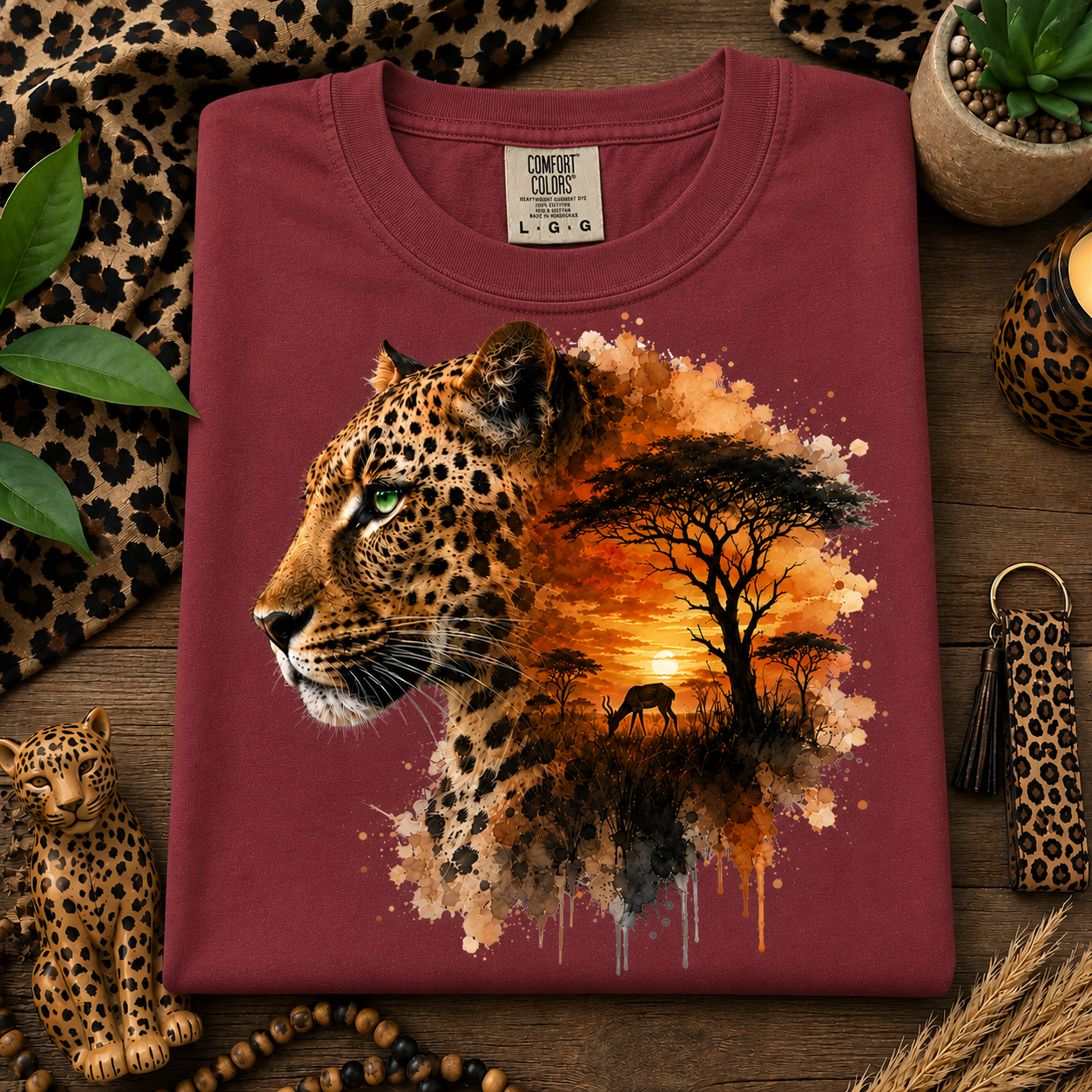 A folded chili colored t-shirt with a graphic print of a leopard and an African savanna sunset, styled on a wooden surface with leopard-print accessories.