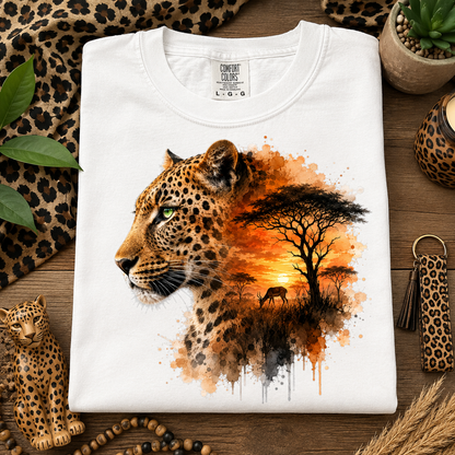 A white short-sleeve T-shirt laid flat on a wooden surface, featuring a large printed graphic of a leopard in profile blending into an African savanna sunset scene with a tree and bird. Leopard-print fabric, a small leopard figurine, leaves, and other styled props surround the shirt.