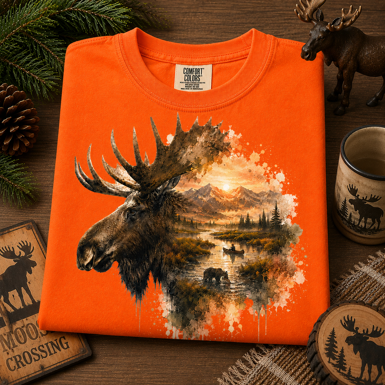 An burnt orange short-sleeve t-shirt with a large graphic print of a moose head and a forest landscape, styled in a flat-lay on a wooden surface with pine branches, a ceramic mug, and moose-themed coasters.
