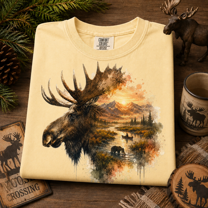 A folded butter colored t-shirt with a detailed graphic print of a moose head merging into a mountain lake landscape at sunset. The shirt is displayed on a rustic wooden surface surrounded by pine branches, a small moose figurine, a ceramic mug, and wooden coasters.