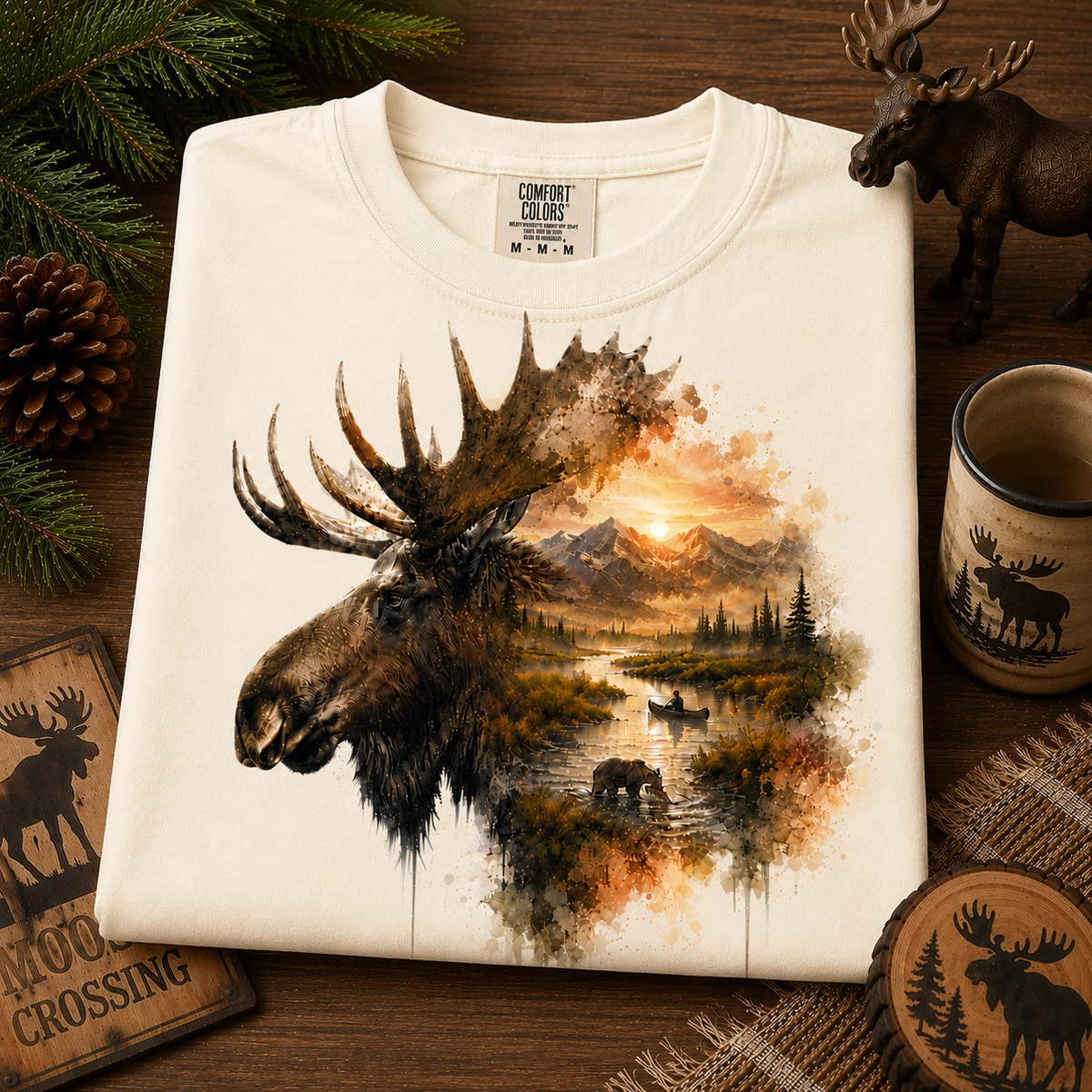 A folded ivory colored Comfort Colors t-shirt with a detailed graphic print of a moose head merging into a forest landscape, displayed on a rustic wooden background with pine needles, a mug, and wooden coasters.