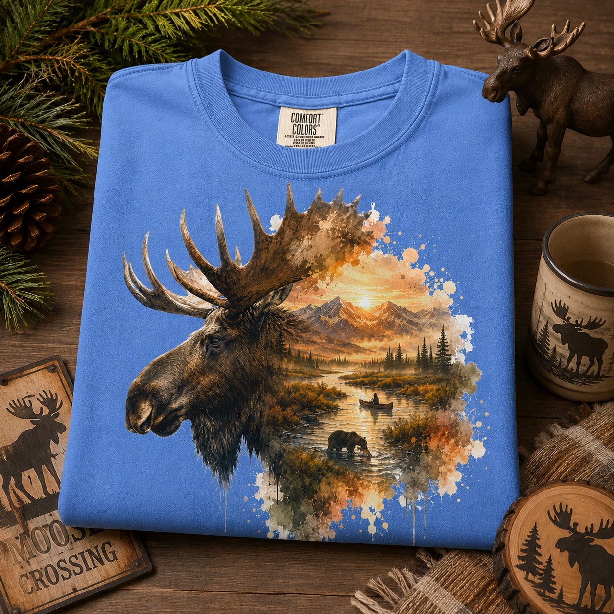 A folded mystic blue t-shirt with a large graphic print of a moose head and a scenic landscape watercolor, styled on a rustic wooden background with pine branches, a mug, and a small moose figurine.